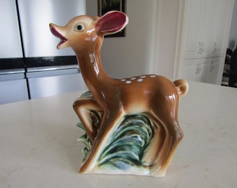 May include: A ceramic deer figurine with a brown body, white spots, and a red-purple inner ear. The deer is standing on a base with green and blue painted foliage. The figurine has an open mouth and a glossy finish.