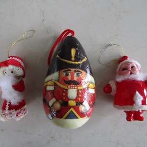 May include: Three Christmas ornaments: a red and white Santa Claus, a red and white nutcracker, and a red and white girl with a white beard.