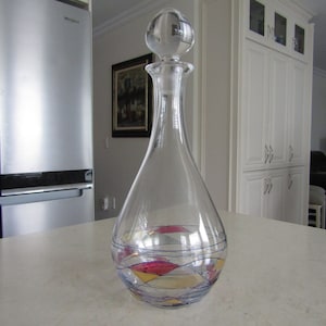May include: A clear glass decanter with a round stopper. The decanter has a geometric design with blue, red, gold, and yellow accents.