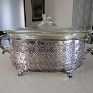 May include: Silver-plated oval casserole dish with a glass lid. The dish has an intricate, ornate design and three feet.