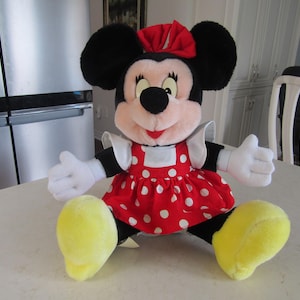 May include: A plush Minnie Mouse doll, featuring black ears, a red bow, and a red dress with white polka dots. The doll has white gloves and yellow shoes, and is sitting on a white surface.