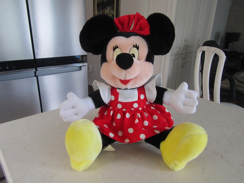 May include: A plush Minnie Mouse doll, featuring black ears, a red bow, and a red dress with white polka dots. The doll has white gloves and yellow shoes, and is sitting on a white surface.