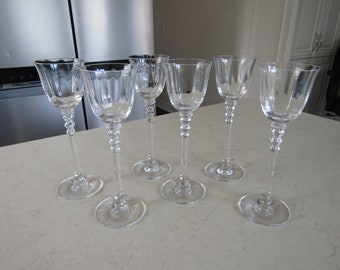 May include: Set of six clear crystal wine glasses. Each glass has a decorative stem with a series of small, round embellishments. The bowls of the glasses have a slightly flared, scalloped rim.
