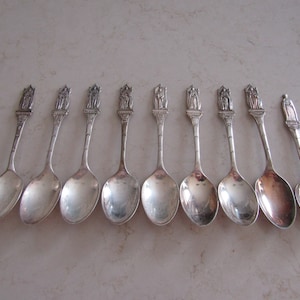 May include: Set of ten silver teaspoons with decorative handles featuring various designs. The spoons are arranged in a row on a light gray surface.