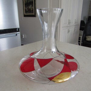 May include: A clear glass decanter with a wide base and a tall neck. The decanter is decorated with a geometric pattern of red, white, and gold.