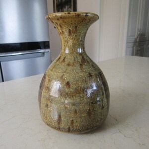May include: A handmade ceramic vase with a speckled, earthy design. The vase has a wide, rounded body that narrows to a flared neck. The colour palette includes shades of olive green, brown, and tan, creating a rustic aesthetic. This decorative vase is suitable for home decor.