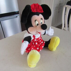 May include: A plush Minnie Mouse doll, featuring black ears, a red bow, and a red dress with white polka dots. The doll has white gloves and yellow shoes, and is sitting on a white surface.