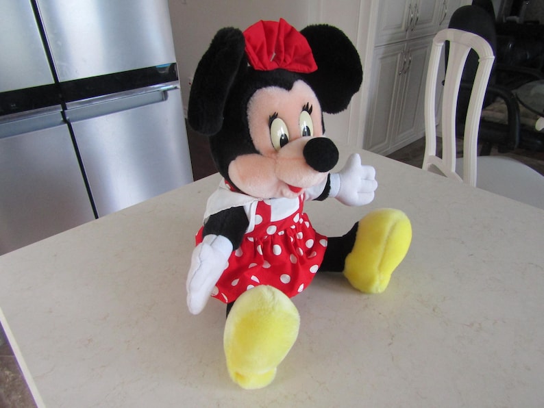 May include: A plush Minnie Mouse doll, featuring black ears, a red bow, and a red dress with white polka dots. The doll has white gloves and yellow shoes, and is sitting on a white surface.