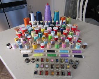 May include: A collection of sewing supplies, including spools of thread in various colors, cones of thread, bobbins, and thread organizers. The spools are in a variety of colors, including red, yellow, blue, and green. The cones are in purple, orange, and blue.