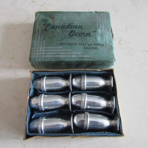 May include: A vintage set of six individual salt and pepper shakers. The box is green and reads "The Canadian Acorn". The shakers are silver-toned and are presented in a blue-lined box. The box is rectangular.