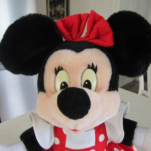 May include: A plush Minnie Mouse doll with large black ears, a red bow, and a red and white polka dot dress. The doll has a black nose, large eyes, and white gloves. The doll is a popular character from Disney.