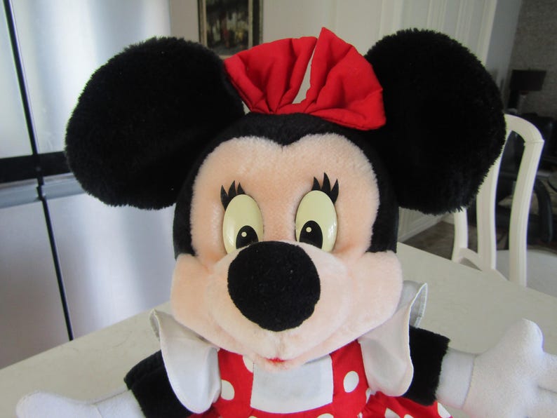 May include: A plush Minnie Mouse doll with large black ears, a red bow, and a red and white polka dot dress. The doll has a black nose, large eyes, and white gloves. The doll is a popular character from Disney.