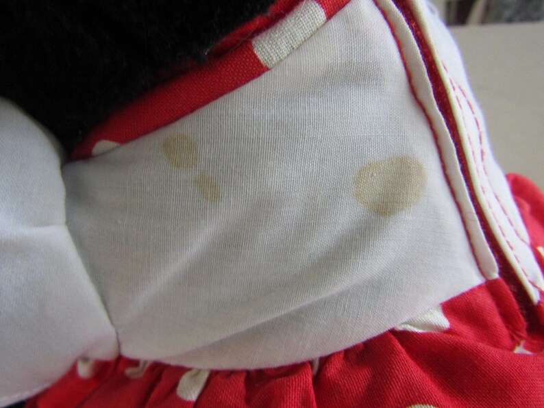 May include: Close-up of a red and white polka dot dress with a white collar. The collar has brown stains. The dress is made of a soft fabric and has a white bow.