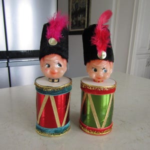 May include: Two vintage Christmas elf drummer decorations. Each elf has a black velvet hat with a pink feather and a smiling face. The drums are red and green with gold trim.