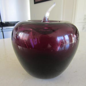 May include: A decorative glass apple in shades of deep purple and black. The apple has a clear glass stem. The smooth, reflective surface of the apple creates a striking visual effect. This decorative piece is suitable for home decor.