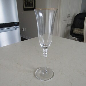 May include: A clear glass champagne flute with a gold rim. The flute has a long stem and a round base.