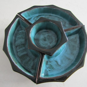 May include: A teal and black ceramic serving dish set. The set includes a central square bowl and four surrounding sections. The teal glaze has a mottled appearance, and the edges are dark brown.