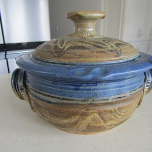 Peut inclure: Un plat à casserole en céramique fait à la main avec un couvercle. Le plat présente une bande bleue au milieu, avec des motifs tourbillonnants bruns et beiges. Il a deux poignées et un bouton sur le couvercle.