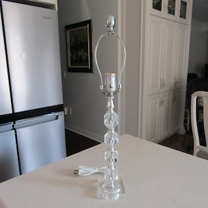 May include: A silver table lamp with a clear crystal base and a white cord. The lamp has a clear glass shade and a silver finial.