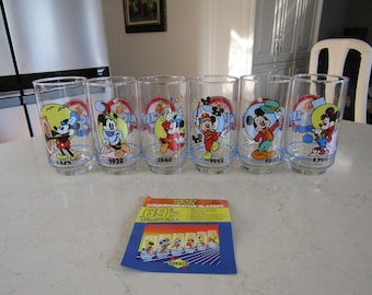 May include: Set of six clear glass tumblers featuring Mickey Mouse in various poses and years. Each glass has a blue rim and a blue band with the text "Mickey Mouse" and the year. The glasses are arranged in a row on a white surface. The text on the card reads "Mickey Mouse Commemorative Glasses 6/9 Collect All 6.  Sondico."