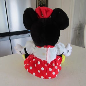 May include: A plush Minnie Mouse doll, viewed from the back, with large black ears and a red bow. She wears a white top, a red skirt with white polka dots, and white gloves. The doll is sitting on a white surface.