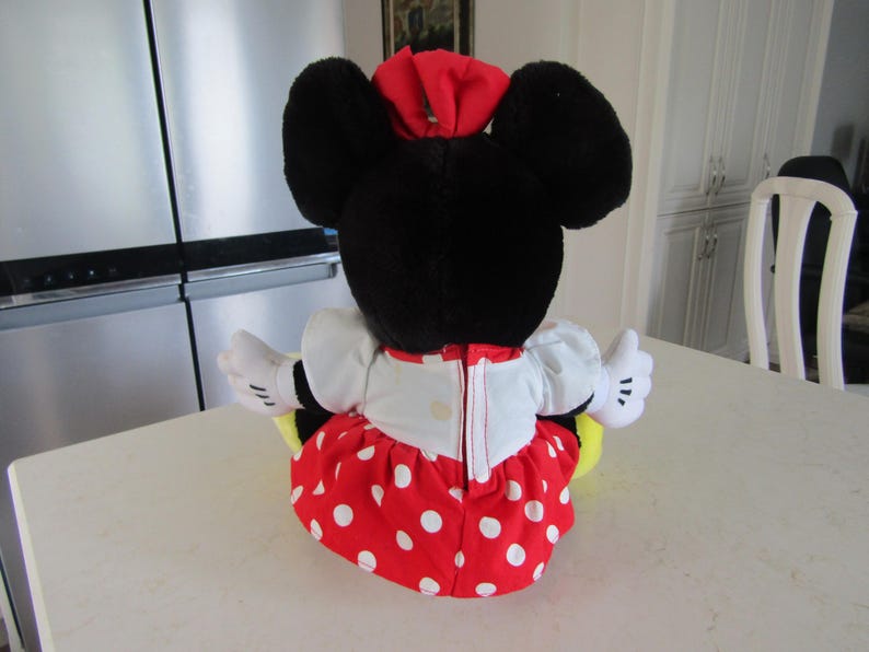May include: A plush Minnie Mouse doll, viewed from the back, with large black ears and a red bow. She wears a white top, a red skirt with white polka dots, and white gloves. The doll is sitting on a white surface.