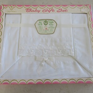May include: A white baby crib set in a pink and green box. The set includes a fitted sheet, a top sheet, and a pillowcase. The top sheet has a lace trim and a satin bow. The box has the text "Baby Gift Set" on the front.