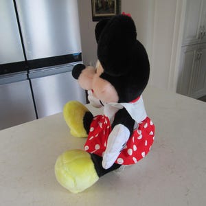 May include: A plush Minnie Mouse doll, featuring a black head with large ears, a red and white polka-dot skirt, and yellow shoes. The doll is sitting on a white surface.