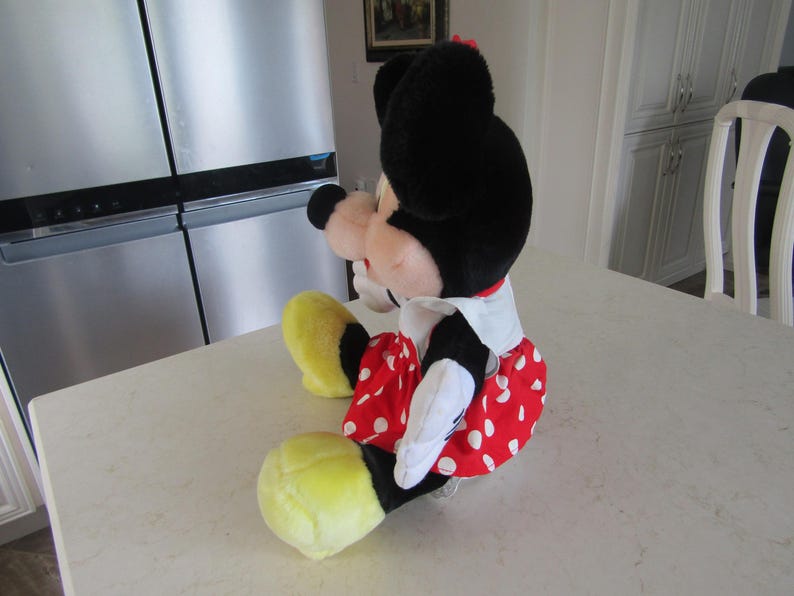 May include: A plush Minnie Mouse doll, featuring a black head with large ears, a red and white polka-dot skirt, and yellow shoes. The doll is sitting on a white surface.