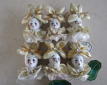 May include: Set of eight porcelain doll ornaments with gold and white accents. Each doll has a white face with painted features and wears a gold and white ruffled collar. The dolls are wearing gold and white hats with bows and are adorned with gold glitter.