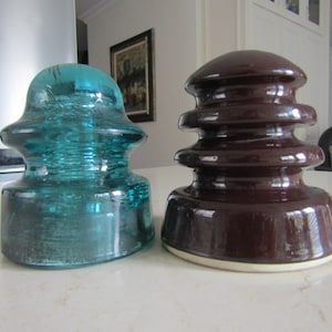 May include: Two antique glass insulators. One is a clear teal colour, and the other is a dark brown. Both have a rounded top and a tiered design. They are sitting on a white surface.
