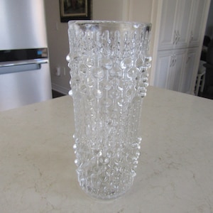 Peut inclure: Un vase en verre transparent avec une surface texturée. Le vase a une forme cylindrique et est décoré d'un motif répétitif de petits points en relief.