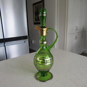 May include: A green glass decanter with a gold-trimmed spout and a silver-striped design. The decanter has a round base and a tall, slender neck.