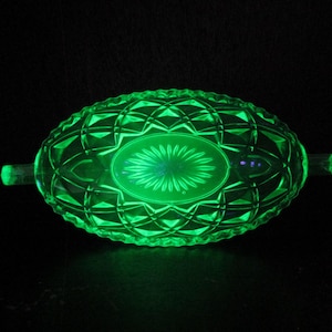 May include: A vibrant green glass dish, featuring a diamond pattern and an oval centre design. The dish has two small handles and glows against a dark backdrop. This vintage piece is likely uranium glass.