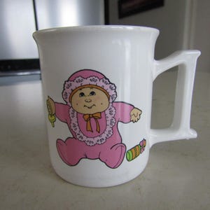 May include: White ceramic mug with a cartoon baby in a pink outfit and bonnet. The baby is holding a rattle and there is a colourful toy on the side. The mug has a large, rectangular handle.
