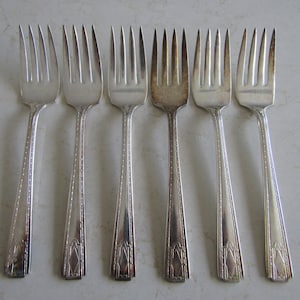 May include: Six silver forks with ornate designs on the handles. The forks are arranged in a row on a white surface.