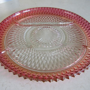 May include: A clear glass serving platter with a divided design. The rim is a vibrant pink with a geometric pattern. The centre features a starburst design. This vintage dish is perfect for serving appetizers or snacks.