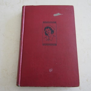 May include: A vintage, red hardcover book with a black and white illustration of a woman in a nurse's cap. The book's title is printed in black above and below the illustration. The cover shows some wear and tear.