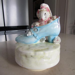 May include: A ceramic figurine featuring a light blue shoe with a high heel, housing a mouse family. The mice are white and gray with pink accents. The shoe sits atop a round, off-white base with green and yellow hues.