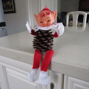May include: A red and white elf doll with a pine cone for a body. The elf is sitting on a white countertop.