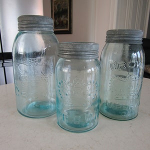 Peut inclure: Trois bocaux Mason vintage en verre bleu avec des couvercles en métal argenté. Les bocaux sont estampillés du mot "CROWN" et d'autres textes. Les bocaux sont de tailles différentes et sont disposés sur une surface blanche.