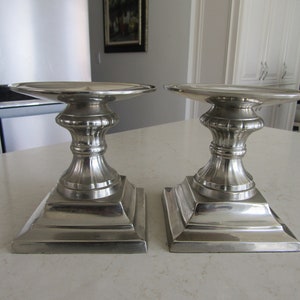 May include: Two silver-colored candle holders with a square base and a decorative, fluted column. The top of each holder is a wide, flat disc. These decorative pieces are suitable for home decor.