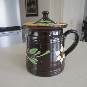 May include: A brown ceramic pitcher with a lid, featuring a white flower with a red center and green leaves. The pitcher has a handle and a spout.
