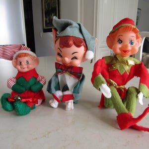 May include: Three vintage Christmas elf figurines. The elves are made of felt and plastic. One elf is wearing a red and white striped hat, a green and red suit, and a bell. Another elf is wearing a green and white hat, a green and red suit, and a plaid bow tie. The third elf is wearing a red hat, a red and green suit, and a gold belt.