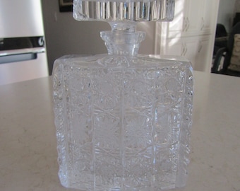 May include: A clear glass decanter with a square body and a faceted stopper. The decanter has a cut glass design with a repeating pattern of flowers.