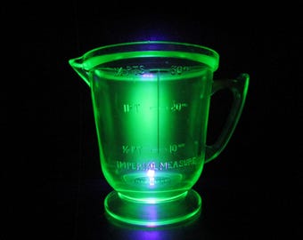 May include: A green glass measuring cup with a handle. The cup has markings for 1/4 pint, 1/2 pint, 1 pint, 10 fl oz, 20 fl oz, and 30 fl oz. The cup is illuminated from below, making it glow.