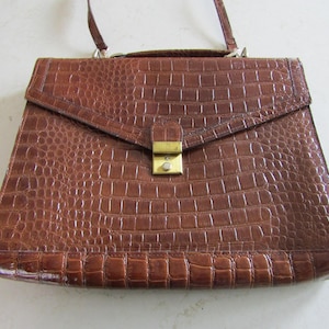 May include: A brown, vintage-style handbag with a textured, crocodile-like pattern. The bag features a flap closure with a gold-colored clasp and a top handle, along with a detachable shoulder strap. The bag's dimensions are approximately 12 inches wide and 9 inches high.