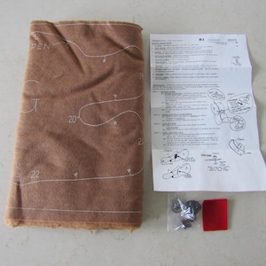 May include: A brown fabric with white lines and numbers printed on it. The fabric is folded in half and there are instructions on a white sheet of paper next to it. There are also small metal pieces and a red fabric square.
