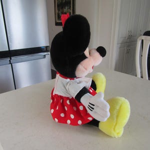 May include: A plush Minnie Mouse doll, seated on a white surface. The doll features a black head with large ears, a red bow, and a red dress with white polka dots. White gloves and yellow shoes complete the classic look.