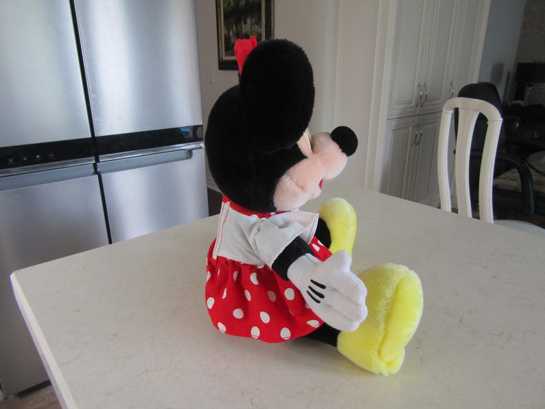 May include: A plush Minnie Mouse doll, seated on a white surface. The doll features a black head with large ears, a red bow, and a red dress with white polka dots. White gloves and yellow shoes complete the classic look.